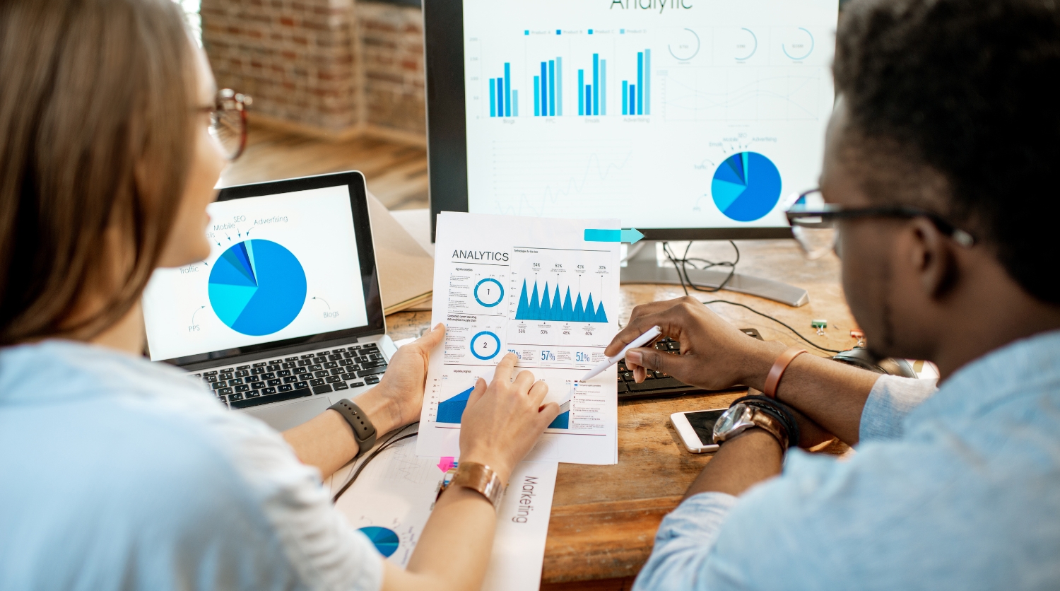 Two automotive marketing professionals reviewing DMS data analytics and pie charts on a computer screen and printed reports.